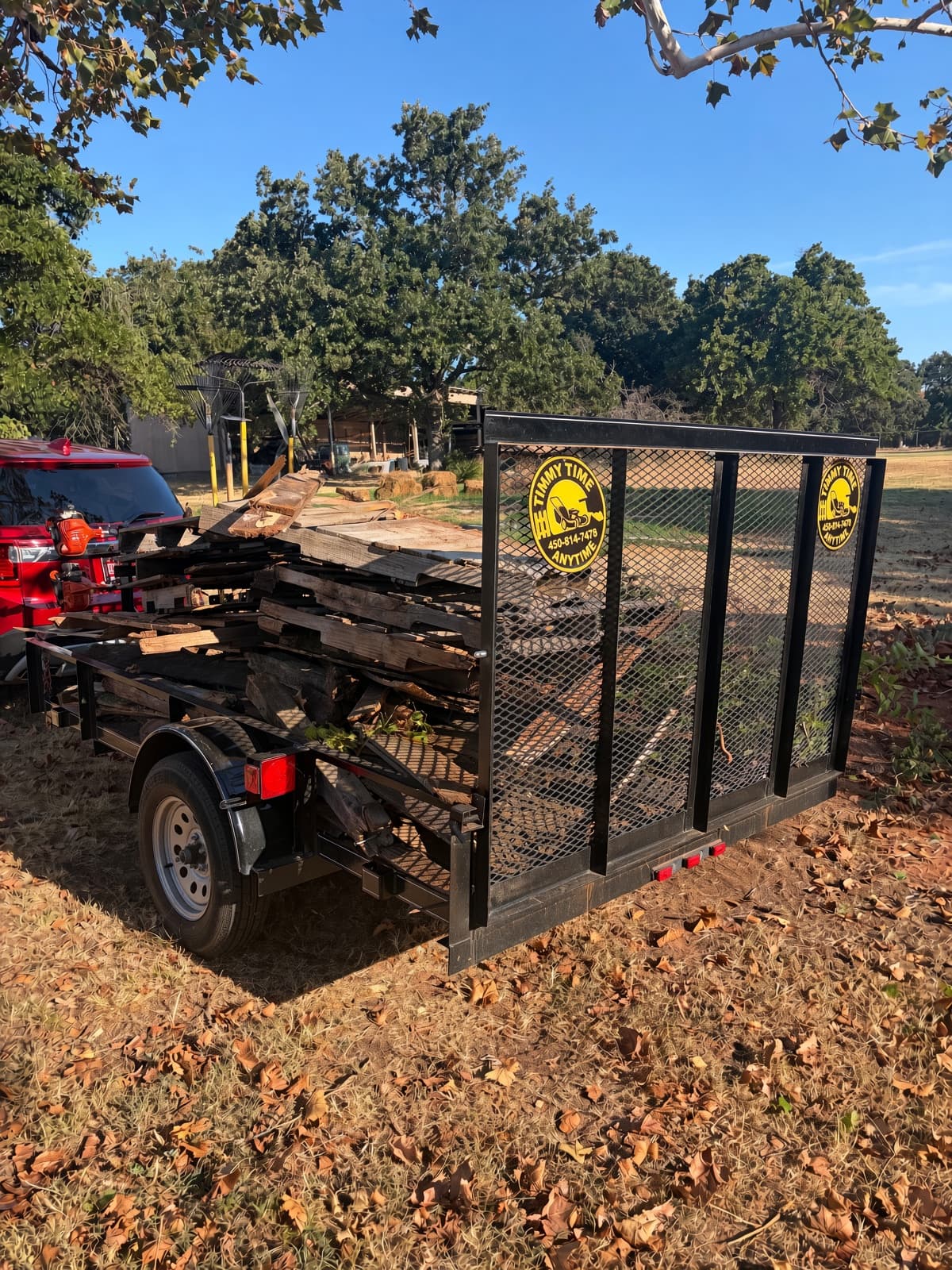 A black utility trailer parked in a sunlit field, heavily loaded with a large pile of weathered scrap wood and broken pallets for hauling.